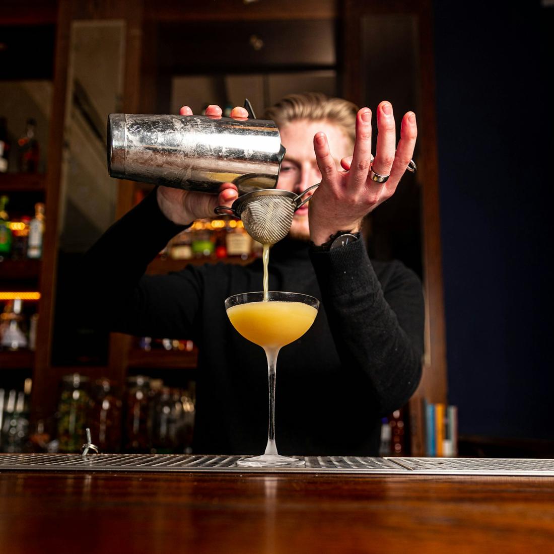 A bartender straining a golden cocktail into a coupe glass