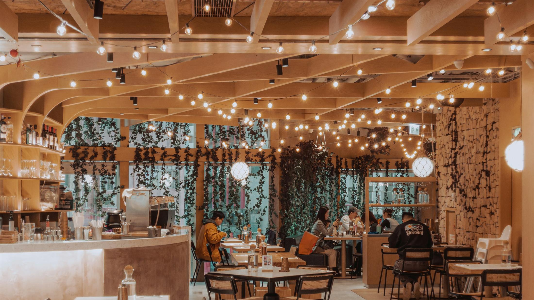 The EMBER & OLIVE dining room in the evening, timber beams hung with string lights above rattan chairs and a curved marble bar