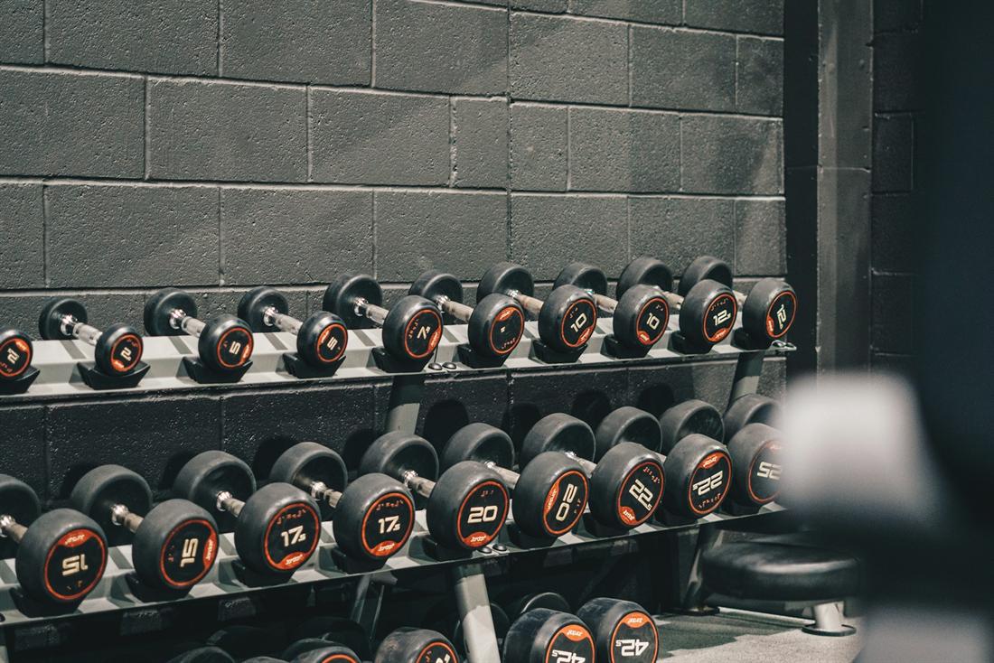 Racks of dumbbells with orange numbers against a grey brick wall