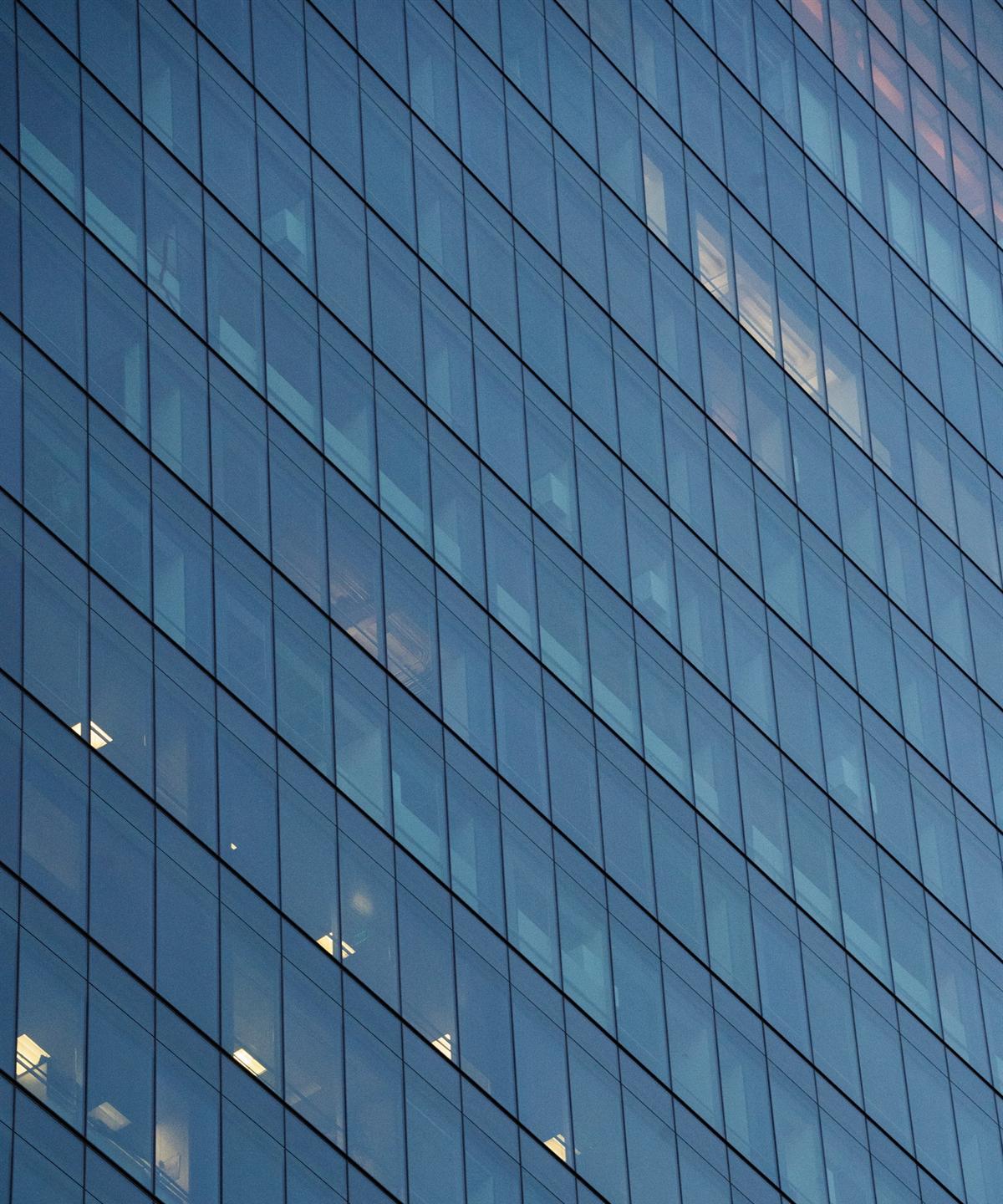 A glass curtain-wall building façade with lit windows reflecting a blue sky