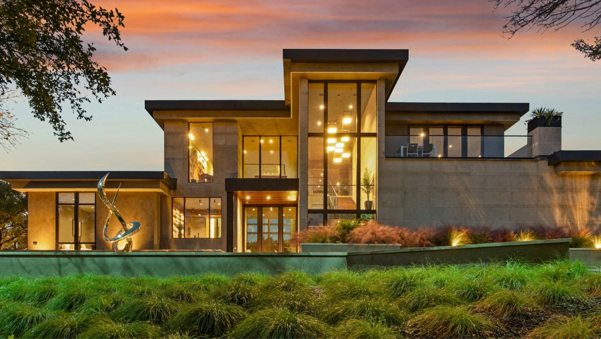 A modern concrete and glass home with tall windows glowing at dusk behind a landscaped garden